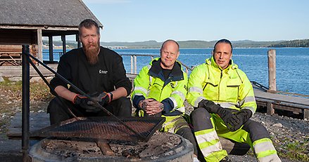 tre personer i arbetskläder sitter vid en grillplats med havet i bakgrunden