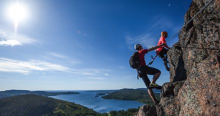 två personer med rep som klättrat uppför ett högt berg. I bakgrunden en vy över en skärgård.