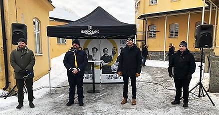 Fyra män på rad utomhus. Vid grinden till riksanstalten, gul stenbyggnad