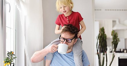 Man med laptop framför sig. Ett barn på axlarna och en kaffekopp i handen.