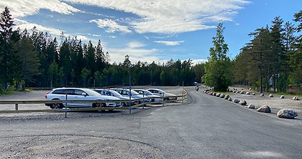 en stor, öppen parkering med några bilar parkerade. Parkeringen omges av skog.