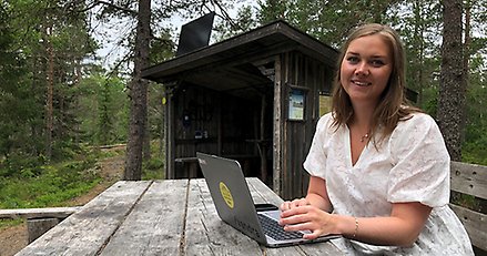 En kvinna sitter med en bärbar dator vid ett utebord. I bakgrunden syns ett vindskydd med en solpanel på taket.