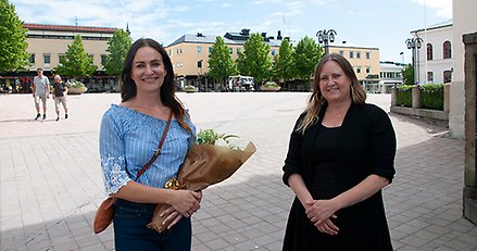 två kvinnor med ett torg i bakgrunden. Den ena har en blombukett i handen 