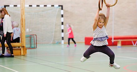 En liten flicka hänger i ett par romerska ringar i en idrottshall. I bakgrunden andra barn och ungdomar.