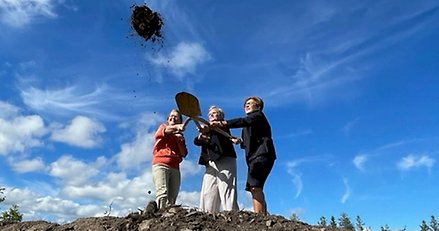 tre kvinnor står på en jordhög och kastar tillsammans jord från en spade med tre handtag