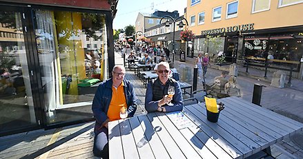 två män sitter vis ett bord på en uteservering i stadsmiljö