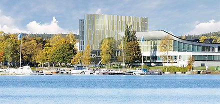 Skissbild på simhallen i Härnösand i förgrunden och ett något högre grått hotell i bakgrunden. I framkant en småbåtshamn.