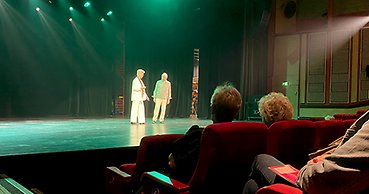 En man och en kvinna visar kläder för en publik i en nedsläckt teatersalong.