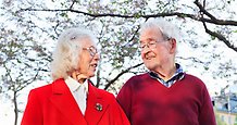 ett äldre par, en kvinna och en man, ser på varandra och ler. I bakgrunden finns ett träd