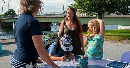 en kvinna med två barn står vid ett bord med en karta på och pratar med en person man ser ryggtavlan på