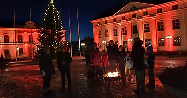 Människor står kring ett eldfat på torget i Härnösand. I bakgrunden residenset och konsthallen upplyst i orange.