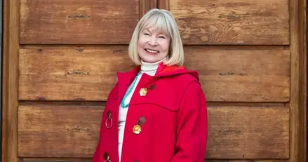 En leende äldre kvinna i röd kappa står vid en träpanel,