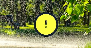 Kraftigt regn på vägbana med grön skog i fonden. En vädervarningssymbol med ett utropstecken på gul botten.