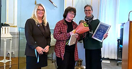 Tre kvinnor uppställda, där en kvinna belönas med ett diplom och blommor.