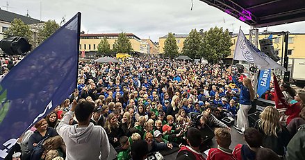 Stor folksamling framför en scen. På scenen står två personer med föreningsflaggor i händerna.