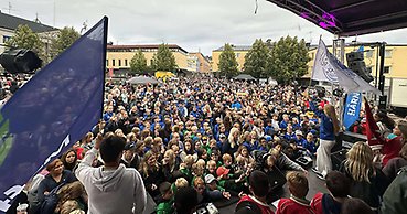 Stor folksamling framför en scen. På scenen står två personer med föreningsflaggor i händerna.