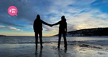 två personer sedda bakifrån står på en strand vid havet och håller varandra i handen