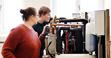 En man och en kvinna tittar nyfiket in i en värmecentral och läser av en mätare. 