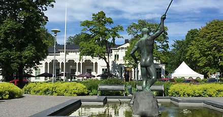 Bronsstaty av naken man med ett ljuster. Statyn står i en grönskande park.