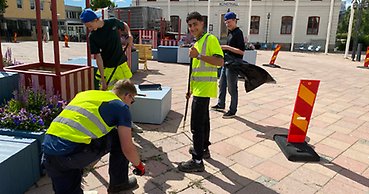 Fyra unga killar med varselvästar rensar ogräs på torget.