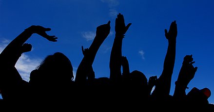 Siluetter av människor i ett publikhav som sträcker upp armar och händer mot en klarblå himmel.