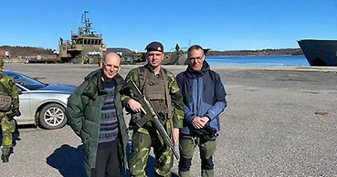 Tre män, varav en är militärklädd. I bakgrunden ett militärfartyg.