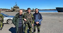 Tre män, varav en är militärklädd. I bakgrunden ett militärfartyg.