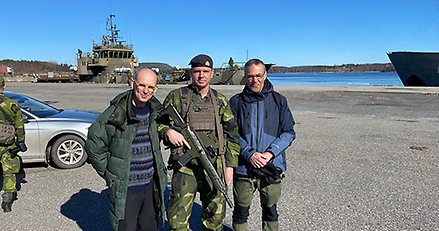 Tre män, varav en är militärklädd. I bakgrunden ett militärfartyg.