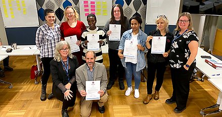 Gruppbild på nio glada personer som står i ett klassrum och stolta visar upp diplom.