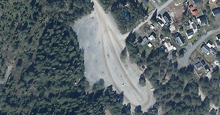 satellitbild med en stor grusplan i mitten omgiven av skog på ena sidan och hus på andra sidan.