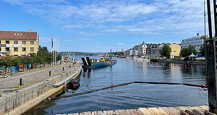 Stor arbetspråm med blått skrov i vattnet intill Nybron. Maskiner är lastade på pråmen.