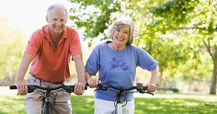 en äldre man och en äldre kvinna sitter på varsin cykel i en grön parkmiljö