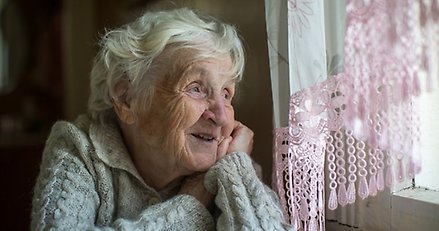 en äldre dam ler och tittar ut genom ett fönster