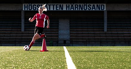 ung kvinna springer med en fotboll på en fotbollsplan. I bakgrunden en skylt där det står Högslätten.