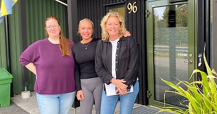 Gruppbild med tre leende kvinnor som står utomhus vid en entrédörr.