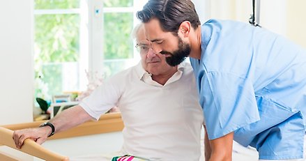 en vårdklädd yngre man hjälper en äldre man  som sitter i en säng