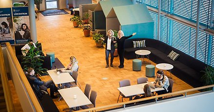 två personer står på en öppen yta och vinkar. Runt om sitter flera personer vid bord och studerar