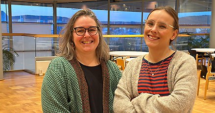 två glada personer med glasögon tittar in i kameran, står i inomhusmiljö