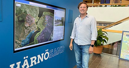 Leende ung man står framför en digital skärm som visar en karta. Under skärmen står textat HÄRNÖSAND.
