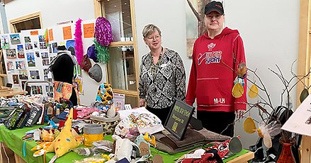 Två kvinnor står bakom ett bord fullt med färggranna textilföremål.