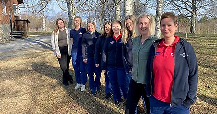 Åtta kvinnor står uppradade utomhus i en björkdunge och tittar leende mot kameran.