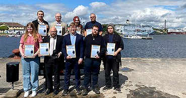 En klass med nio vuxenelever poserar utomhus med sina diplom. I bakgrunden syns ett kryssningsfartyg. 