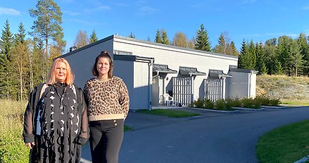 Två kvinnor står utomhus framför en låg byggnad med grå träpanel.