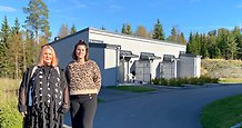 Två kvinnor står utomhus framför en låg byggnad med grå träpanel.