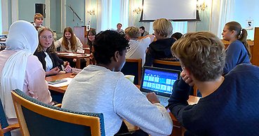 Högstadieelever diskuterar och planerar i en stor möteslokal.