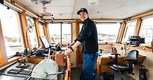 en ung man står vid rodret i en styrhytt på ett stort fartyg