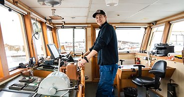 en ung man står vid rodret i en styrhytt på ett stort fartyg