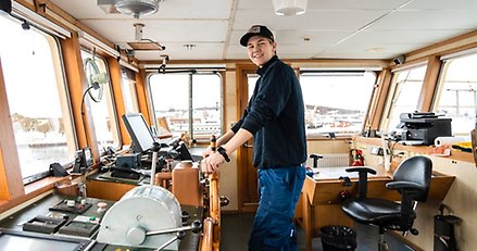 en ung man står vid rodret i en styrhytt på ett stort fartyg