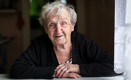 en äldre kvinna sitter med händerna framför sig. Hon ler.