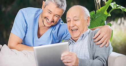 Bild på en manlig omvårdnadspersonal och en äldre man som tittar på en surfplatta